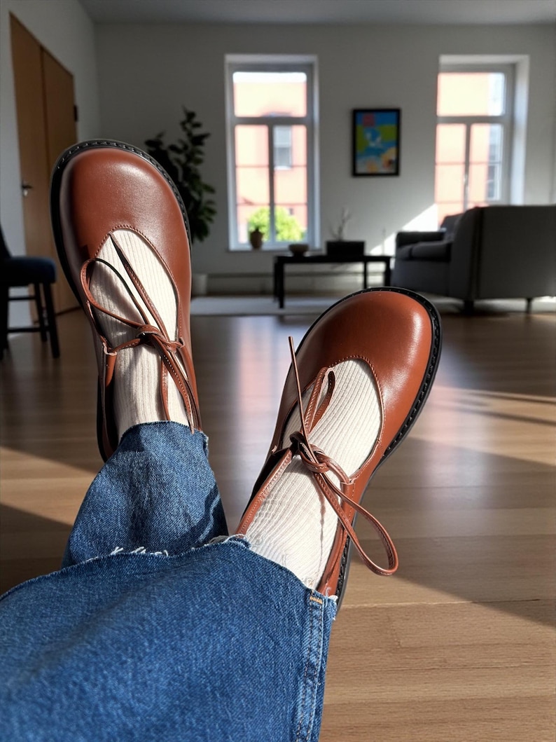 Op de afbeelding: Bruine leren Mary Jane-schoenen met een ronde neus en enkelbandjes. De schoenen worden gedragen met witte sokken en een blauwe spijkerbroek. De schoenen zijn ongeveer 23 cm lang.