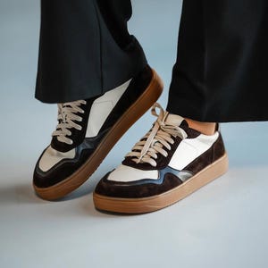 May include: A pair of stylish sneakers with a brown sole, white and brown suede panels, and beige laces. The shoes are worn with black pants, creating a modern, casual look.