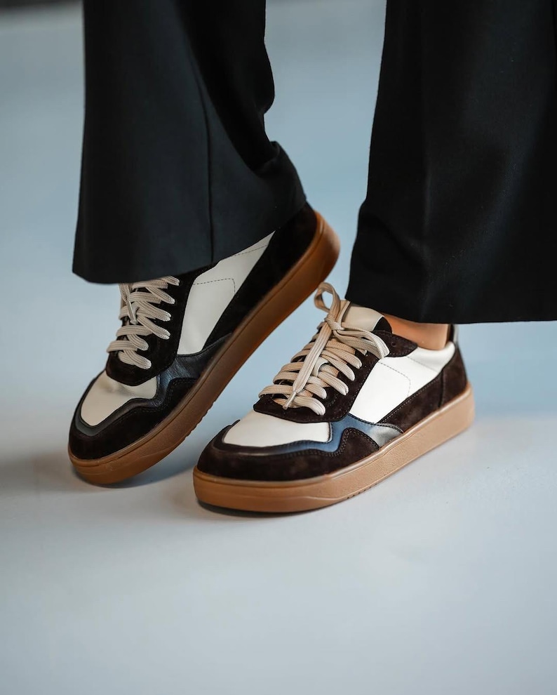 May include: A pair of stylish sneakers with a brown sole, white and brown suede panels, and beige laces. The shoes are worn with black pants, creating a modern, casual look.