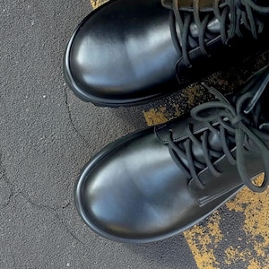 May include: A pair of black leather combat boots with rounded toes and black laces. The boots are on a surface with black asphalt and yellow painted lines. The boots have a classic, sturdy design.