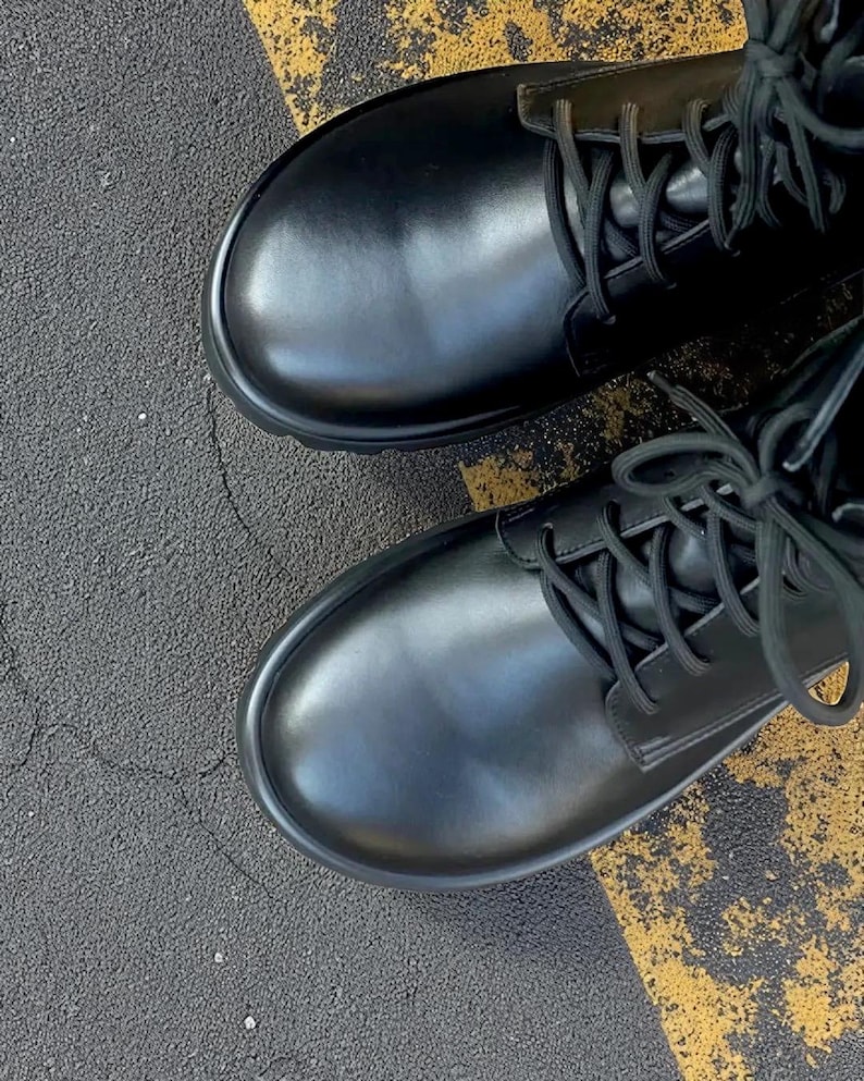May include: A pair of black leather combat boots with rounded toes and black laces. The boots are on a surface with black asphalt and yellow painted lines. The boots have a classic, sturdy design.