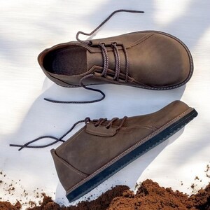 May include: A pair of brown leather shoes with dark brown laces. The shoes have a rounded toe and a dark sole. The shoes are displayed on a white surface with a pile of brown material in the foreground.