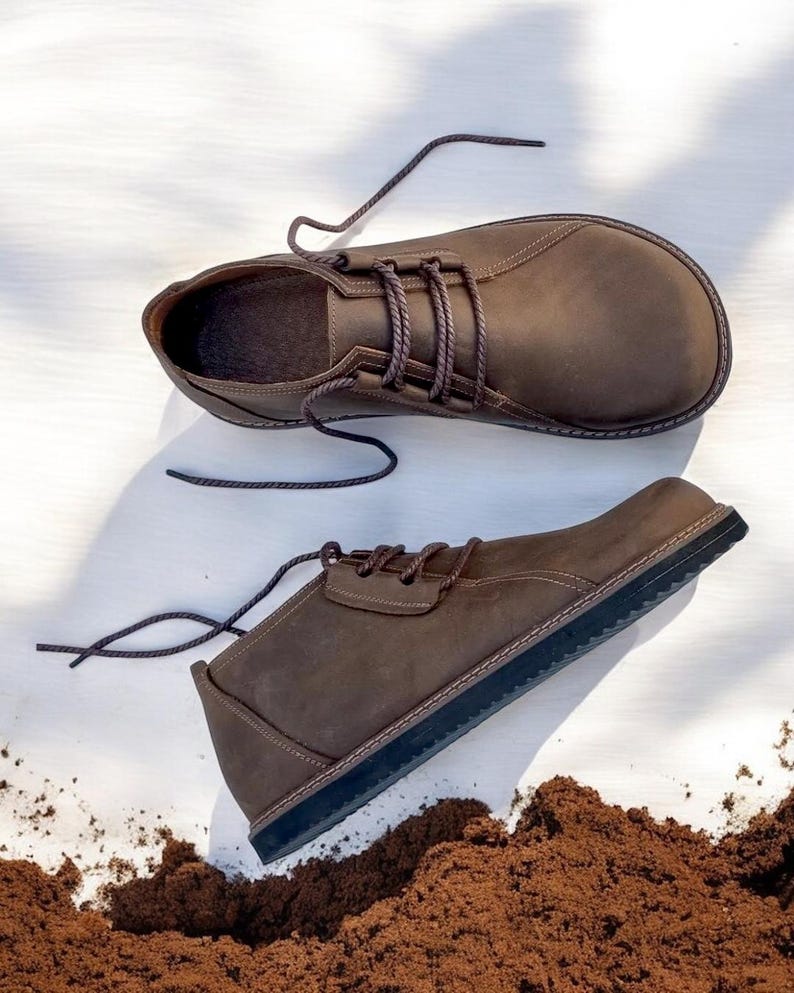 May include: A pair of brown leather shoes with dark brown laces. The shoes have a rounded toe and a dark sole. The shoes are displayed on a white surface with a pile of brown material in the foreground.