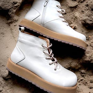 May include: A pair of white leather ankle boots with light brown soles and laces. One boot has a zipper on the side. The boots are set on a sandy surface.