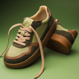 May include: A pair of green and brown sneakers with tan laces. The sneakers have a brown sole and a mix of green and brown panels. The laces are long and hang down. The shoes are on a green background.