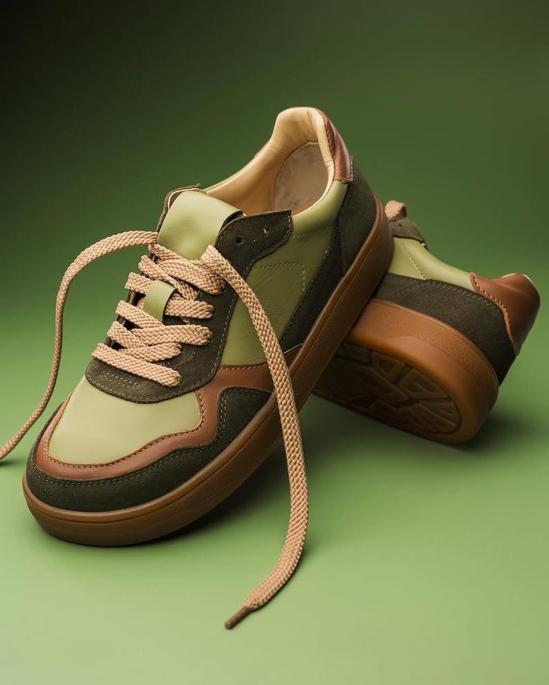 May include: A pair of green and brown sneakers with tan laces. The sneakers have a brown sole and a mix of green and brown panels. The laces are long and hang down. The shoes are on a green background.