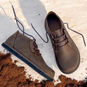 May include: A pair of brown leather chukka boots with dark brown laces. The boots have a rounded toe and a dark sole. The boots are placed on a light-colored surface with a scattering of brown material.