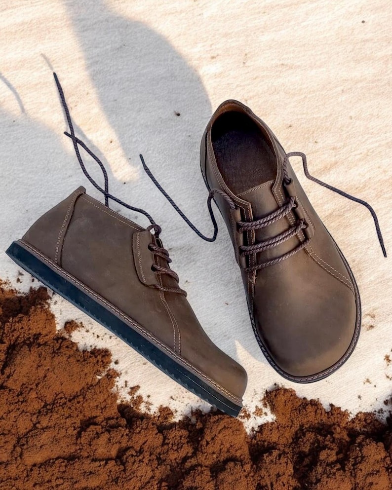 May include: A pair of brown leather chukka boots with dark brown laces. The boots have a rounded toe and a dark sole. The boots are placed on a light-colored surface with a scattering of brown material.