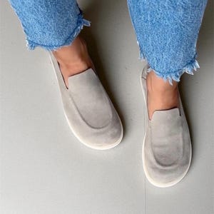 Op de afbeelding: Een paar lichtgrijze su&egrave;de instappers met witte zolen. De schoenen hebben een ronde neus en worden gedragen met een blauwe denim jeans met gerafelde randen. De schoenen zijn een neutrale kleur en passen bij veel outfits.