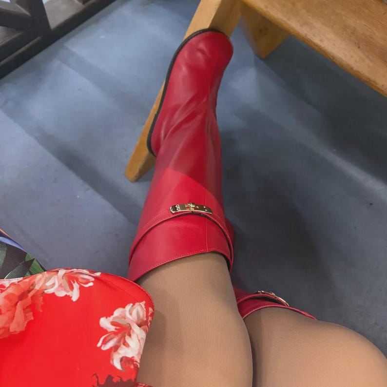 May include: A pair of bright red knee-high boots featuring a smooth finish and a decorative gold-toned metal buckle strap across the upper shaft. The boots are worn with sheer neutral-toned hosiery, paired with a red fabric featuring a white floral print.