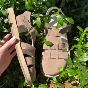 May include: A pair of tan suede sandals with multiple straps and adjustable buckles. The sandals have a thick, light-colored sole and are set against a backdrop of green foliage. The sandals are designed for comfort and style.