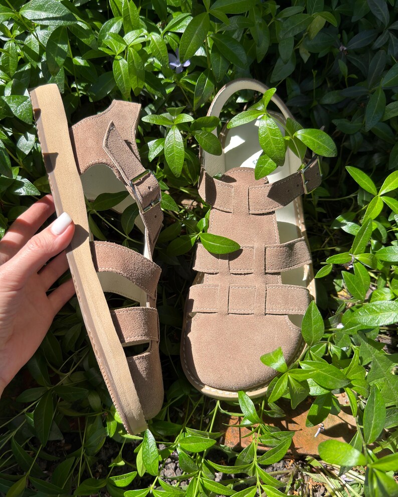 May include: A pair of tan suede sandals with multiple straps and adjustable buckles. The sandals have a thick, light-colored sole and are set against a backdrop of green foliage. The sandals are designed for comfort and style.