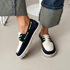 以下が含まれることがあります： 白、ネイビーブルー、グリーンのボートシューズ。白い靴紐が付いています。靴は厚いライトグレーのソールで、革とスエードで作られています。ジーンズに合わせて履かれています。