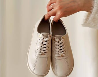 May include: A pair of off-white leather sneakers with matching laces and gold-colored accents. The shoes have a rounded toe and a stitched design. The sneakers are held up by a hand.