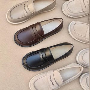 Può includere: Un assortimento di mocassini in pelle in vari colori, tra cui beige, marrone e nero. Le scarpe hanno un design classico con una fascia sul collo del piede e una punta arrotondata. I mocassini sono disposti su una superficie dai toni neutri.