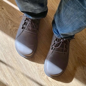 May include: A pair of brown leather shoes with matching laces. The shoes have a rounded toe and are worn with blue jeans. The shoes are on a wooden floor.