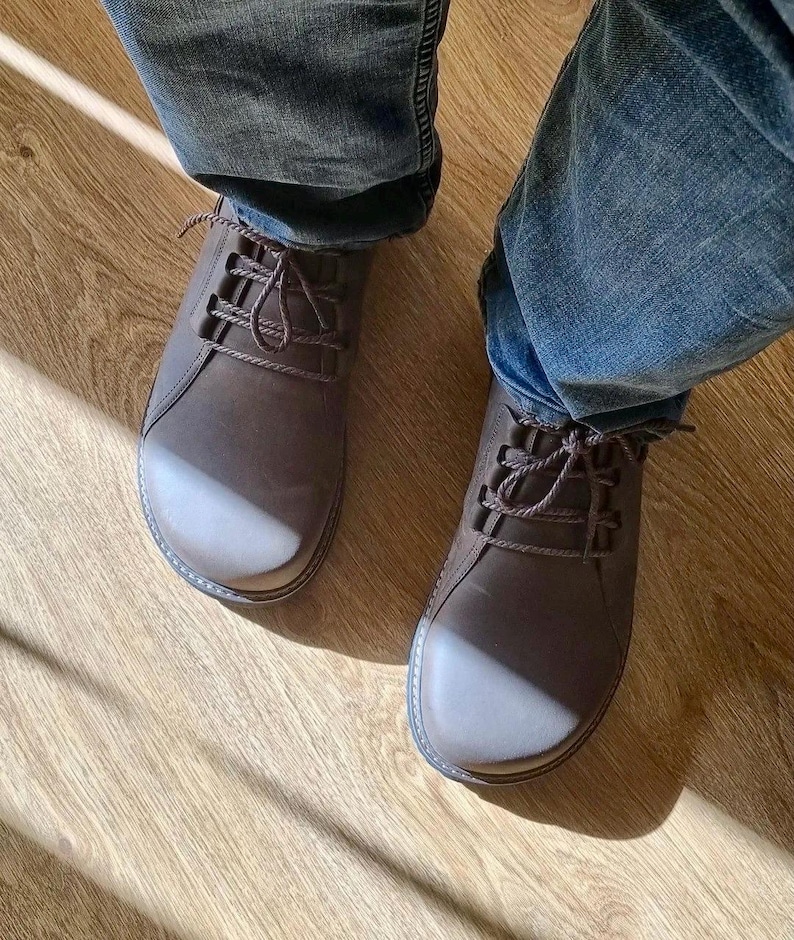 May include: A pair of brown leather shoes with matching laces. The shoes have a rounded toe and are worn with blue jeans. The shoes are on a wooden floor.