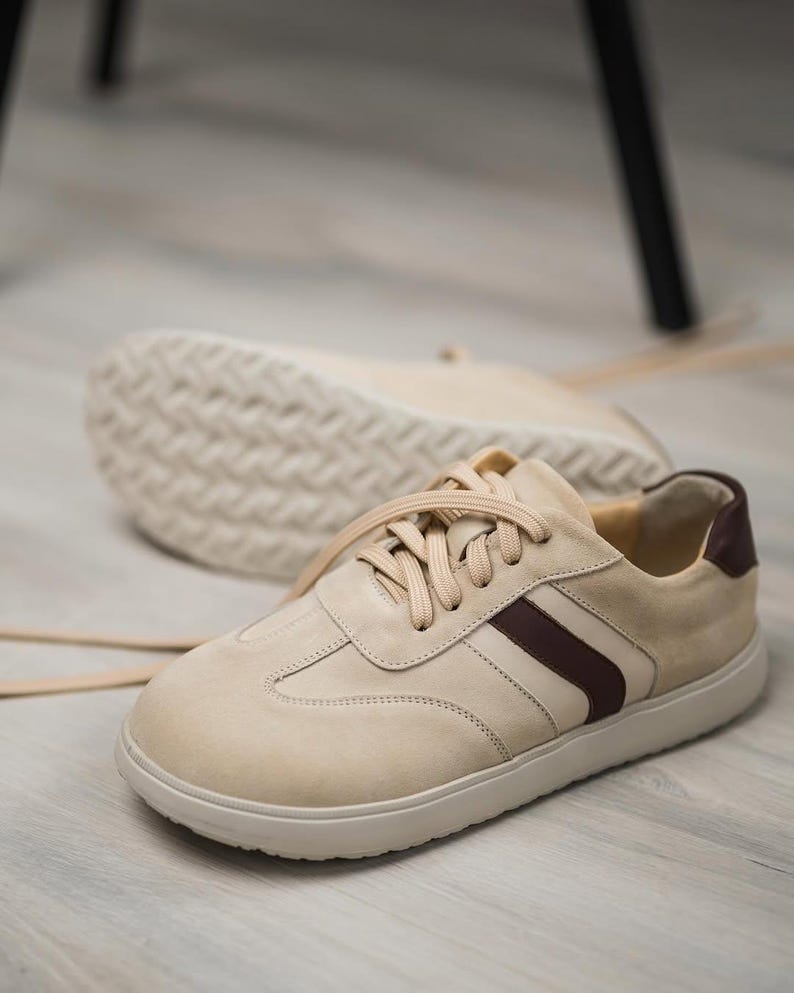 May include: A pair of beige suede sneakers with a brown stripe and white laces. The sneakers have a white rubber sole.