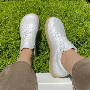 May include: A pair of white leather sneakers with a rounded toe and beige soles. The sneakers have white laces and are set against a backdrop of green grass.