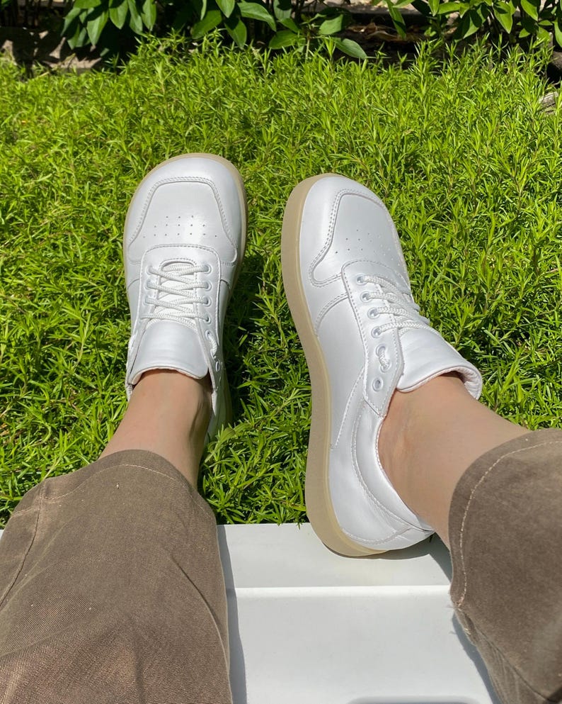 May include: A pair of white leather sneakers with a rounded toe and beige soles. The sneakers have white laces and are set against a backdrop of green grass.