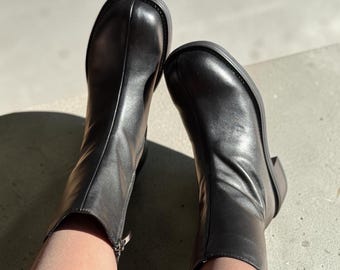 May include: A pair of black ankle boots featuring a smooth finish, a rounded toe box with a central seam detail, and a side zipper closure with a silver-toned pull. The boots have a low block heel and a thick, durable sole construction.