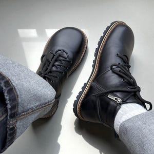 Puede incluir: Botas de cuero negro con diseño de cordones y cremallera lateral. Las botas tienen punta redondeada y ribete marrón alrededor de la suela. Las suelas son negras con un patrón texturizado.