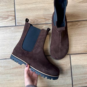 May include: Two brown suede Chelsea boots with elastic side panels and lug soles. The boots have a pull tab at the back and a dark blue and tan trim on the sole. The boots are displayed on a light brown wooden surface.