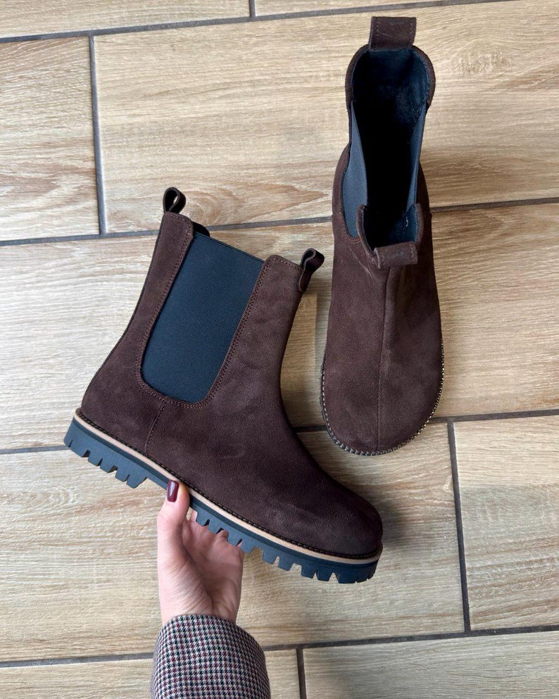 May include: Two brown suede Chelsea boots with elastic side panels and lug soles. The boots have a pull tab at the back and a dark blue and tan trim on the sole. The boots are displayed on a light brown wooden surface.