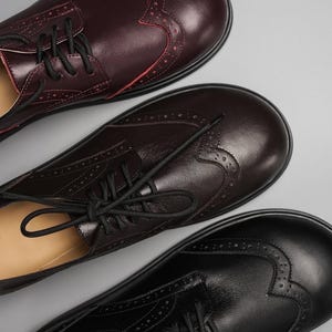 Könnte beinhalten: Drei Paar Oxford-Schuhe aus Leder in Burgunderrot, Braun und Schwarz. Jeder Schuh hat dekorative Perforationen und schwarze Schnürsenkel. Die Schuhe sind diagonal angeordnet und zeigen ihr Design und ihre Farbvariationen.