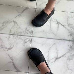May include: Black leather loafers with a rounded toe and a slip-on design. The shoes have a thick sole and are worn with denim jeans. The image is taken from a high angle, showing the shoes on a marble tile floor.