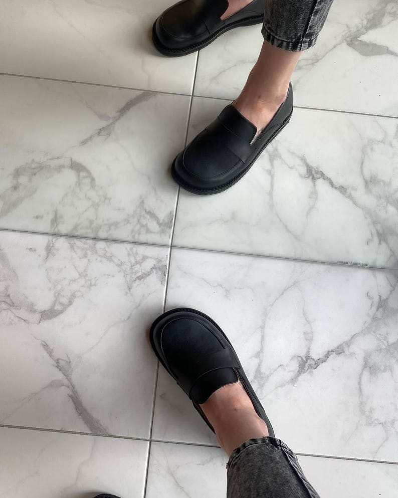 May include: Black leather loafers with a rounded toe and a slip-on design. The shoes have a thick sole and are worn with denim jeans. The image is taken from a high angle, showing the shoes on a marble tile floor.