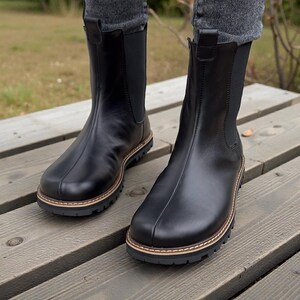 May include: A pair of black leather Chelsea boots with a round toe and a lug sole. The boots have a pull tab at the back and elastic side panels. The soles have a brown and tan trim. The boots are worn with gray jeans.