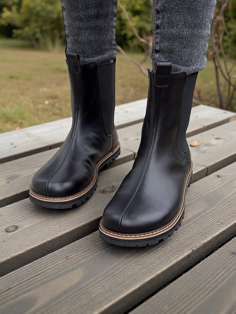 May include: A pair of black leather Chelsea boots with a round toe and a lug sole. The boots have a pull tab at the back and elastic side panels. The soles have a brown and tan trim. The boots are worn with gray jeans.