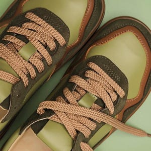 May include: A pair of olive green sneakers with brown accents and tan laces. The shoes have a suede and leather design, with a light green sole and a brown trim. The laces are thick and woven. The sneakers are set against a light green background.