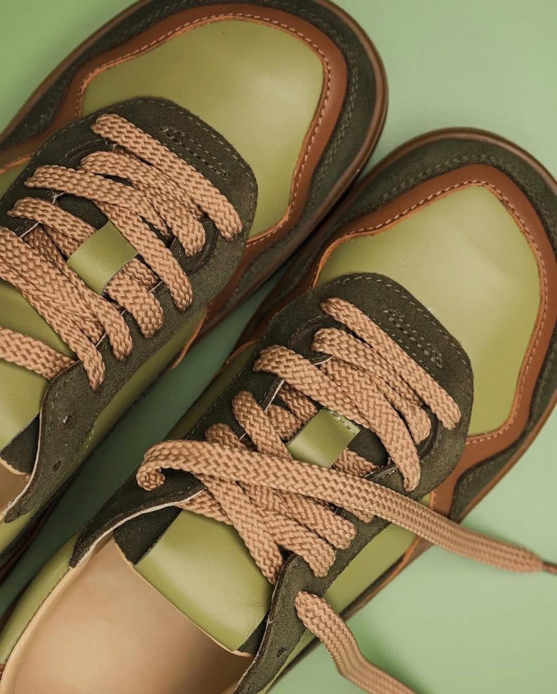 May include: A pair of olive green sneakers with brown accents and tan laces. The shoes have a suede and leather design, with a light green sole and a brown trim. The laces are thick and woven. The sneakers are set against a light green background.