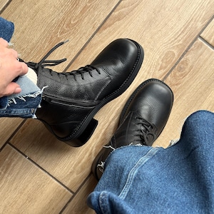 Puede incluir: Un par de botas de cuero negro con cordones, acabado texturizado, puntera redondeada y cierre de cremallera lateral. Las botas cuentan con cordones negros, una suela gruesa negra y un tacón bajo, combinadas con vaqueros azules de dobladillo deshilachado.