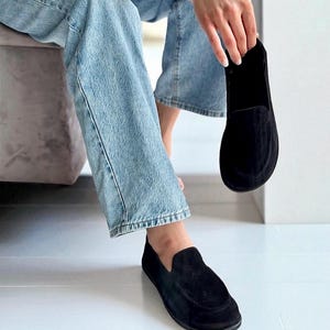 Op de afbeelding: Zwarte su&egrave;de instappers. De schoenen hebben een ronde neus en een laag profiel. E&eacute;n schoen zit aan de voet en de andere wordt omhoog gehouden. De schoenen zijn een klassieke stijl.