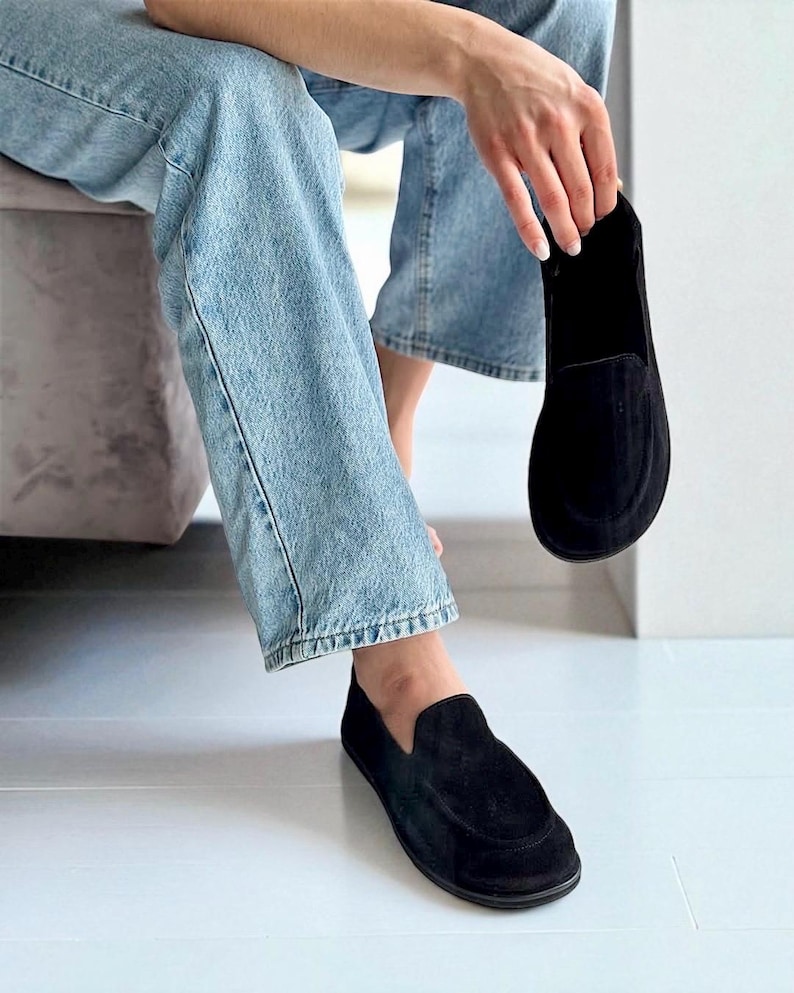 Op de afbeelding: Zwarte su&egrave;de instappers. De schoenen hebben een ronde neus en een laag profiel. E&eacute;n schoen zit aan de voet en de andere wordt omhoog gehouden. De schoenen zijn een klassieke stijl.