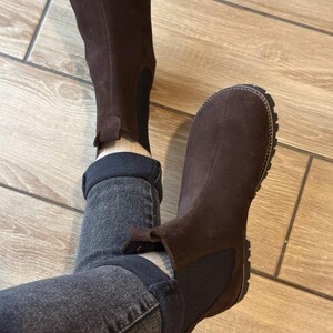 May include: Brown suede Chelsea boots with a dark brown elastic side panel and a lug sole. The boots are worn with dark gray denim jeans. The boots have a pull tab at the back.