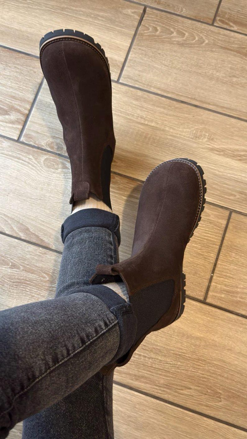 May include: Brown suede Chelsea boots with a dark brown elastic side panel and a lug sole. The boots are worn with dark gray denim jeans. The boots have a pull tab at the back.