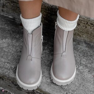 May include: A pair of light gray ankle boots with white soles and zippers. The boots have a front zipper closure and are worn with white ribbed socks. The boots are made of a smooth material and have a chunky sole.