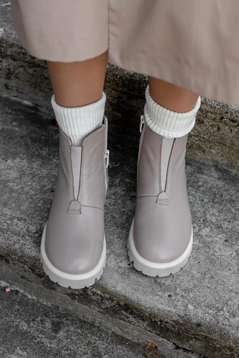 May include: A pair of light gray ankle boots with white soles and zippers. The boots have a front zipper closure and are worn with white ribbed socks. The boots are made of a smooth material and have a chunky sole.