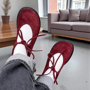 Op de afbeelding: Bordeaux Mary Jane schoenen met een ronde neus en een zwarte zool. De schoenen hebben een witte inzet en zijn vastgebonden met bordeauxrode veters. De schoenen worden gedragen met witte sokken en een grijze jeans.
