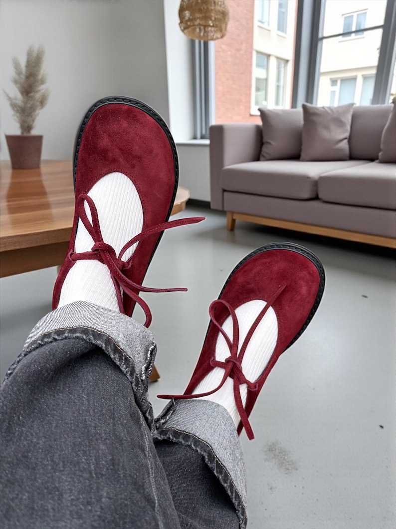 Op de afbeelding: Bordeaux Mary Jane schoenen met een ronde neus en een zwarte zool. De schoenen hebben een witte inzet en zijn vastgebonden met bordeauxrode veters. De schoenen worden gedragen met witte sokken en een grijze jeans.