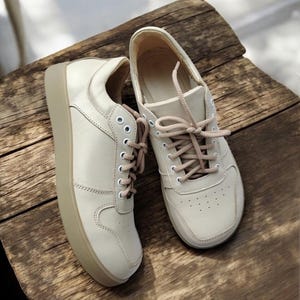 May include: A pair of off-white sneakers with beige soles and matching laces. The shoes are made of a smooth material and have a classic, low-top design. The sneakers are displayed on a rustic wooden surface.