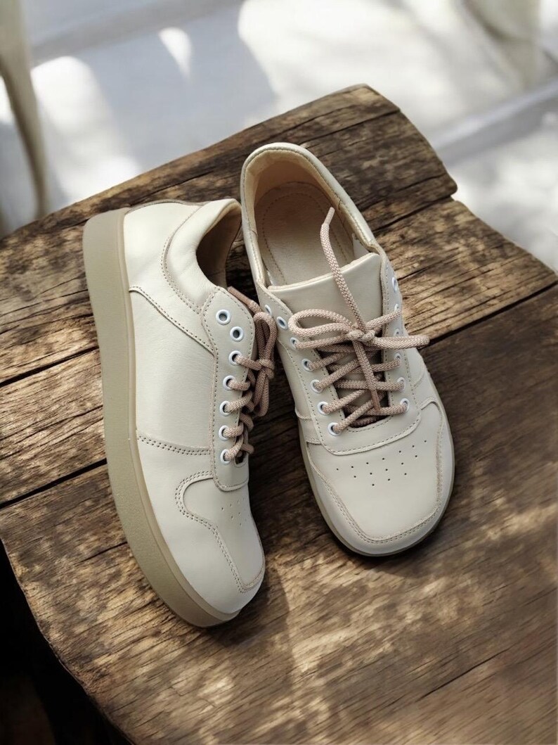 May include: A pair of off-white sneakers with beige soles and matching laces. The shoes are made of a smooth material and have a classic, low-top design. The sneakers are displayed on a rustic wooden surface.