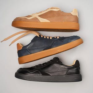 May include: Three pairs of sneakers in a studio shot. The top pair is tan and beige, the middle pair is navy blue, and the bottom pair is black. All have brown soles and laces.
