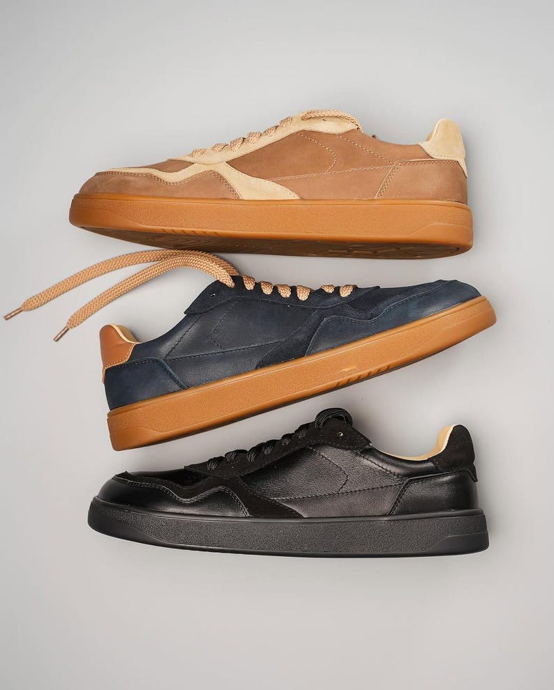 May include: Three pairs of sneakers in a studio shot. The top pair is tan and beige, the middle pair is navy blue, and the bottom pair is black. All have brown soles and laces.