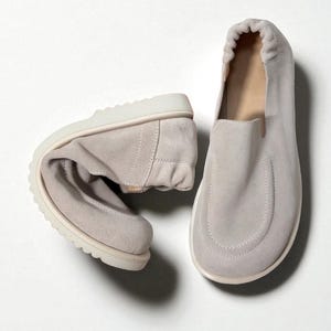 Op de afbeelding: Twee lichtgrijze su&egrave;de instappers. E&eacute;n schoen is dubbelgevouwen, wat de flexibiliteit aantoont. De andere staat rechtop en toont een ronde neus en een cr&egrave;mekleurige zool. De schoenen hebben een minimalistisch, modern ontwerp.