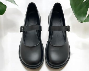May include: A pair of black Mary Jane shoes with buckle straps. The shoes have a rounded toe and a thick sole. The background is white with green leaves.